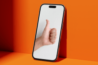 Smartfon na pomarańczowym tle, na którego ekranie widoczna jest dłoń pokazująca kciuk w górę