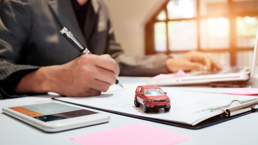 Osoba siedząca przy biurku z długopisem w ręku obok modelu czerwonego samochodu, smartfona i dokumentów, pracująca nad ubezpieczeniem auta.