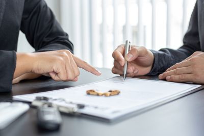 Osoba podpisuje dokument, prawdopodobnie umowę, z drugą osobą wskazującą na miejsce podpisu, z kluczami samochodowymi na stole.