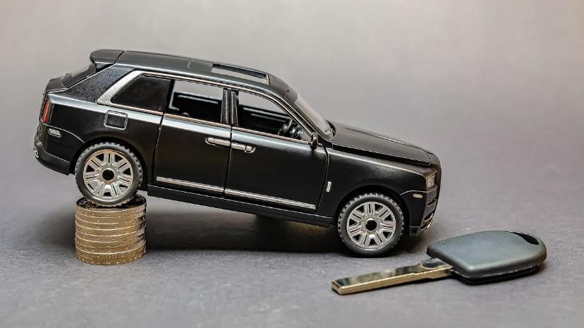 Model luksusowego SUV-a balansujący na stosie monet, symbolizujący utratę wartości samochodów, obok kluczyka samochodowego