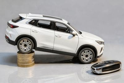 Model białego SUV-a balansujący na stosie monet, symbolizujący utratę wartości samochodów, obok kluczyka samochodowego